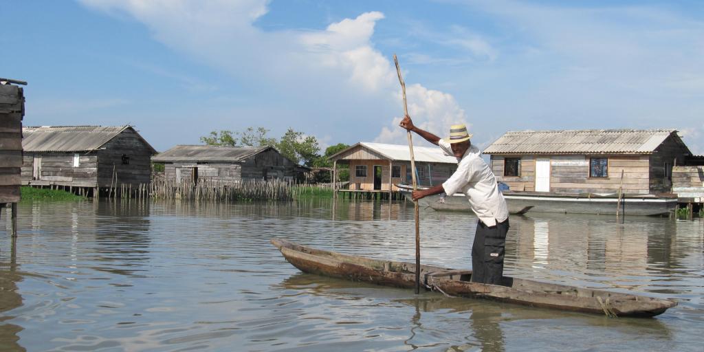 Hombre en una canoa remando con ayuda de una vara, detrás de él aparecen casas flotantes.