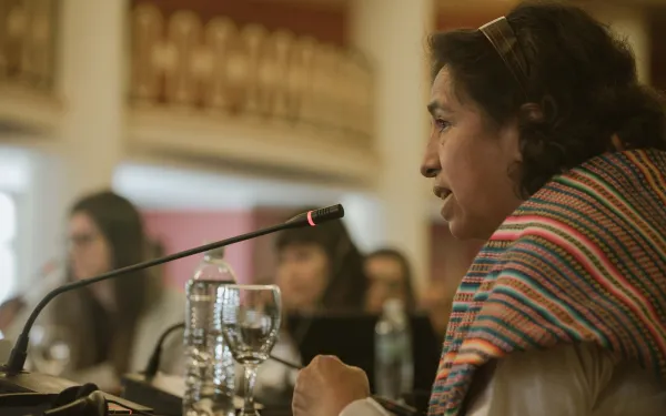 una mujer, envuelta en un chaleco tradicional, declara ante un micrófono