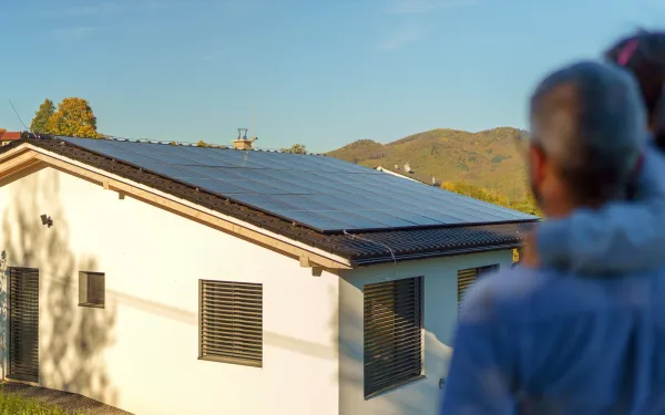 Vista trasera de un padre con su hija en brazos y mirando su casa con paneles solares instalados. 